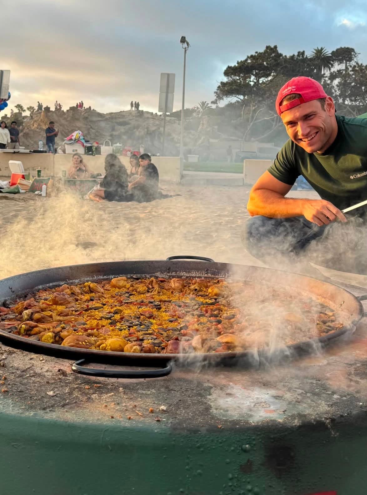 Pau, the chef behind Pauella, smiling next to a steaming paella pan on the beach